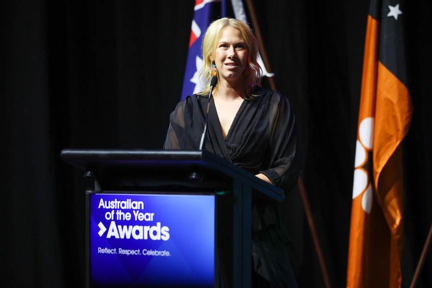 Blonde-haired woman stands at lectern giving a speech with Australian flag behind her.