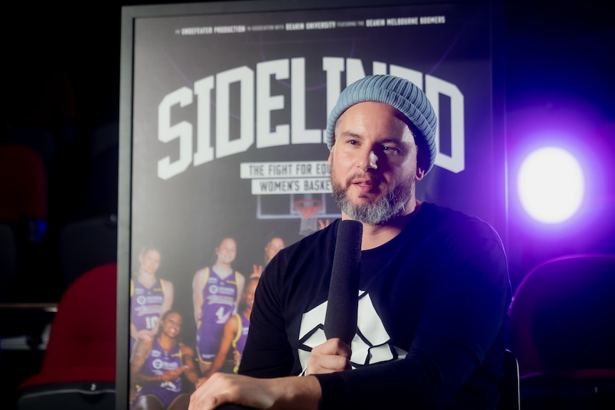 Filmmaker Adam McKay is sitting, holding a microphone and talking, a 'Sidelined' film poster is behind him.