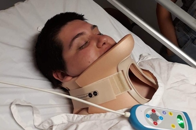A young woman with short hair lies on what looks like a hospital bed, in a neck brace.