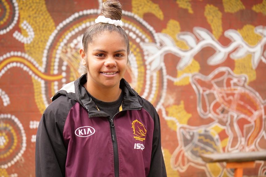 a young woman smiling at the camera, hair in a top knot, wearing a broncos jacket, colourful background.,