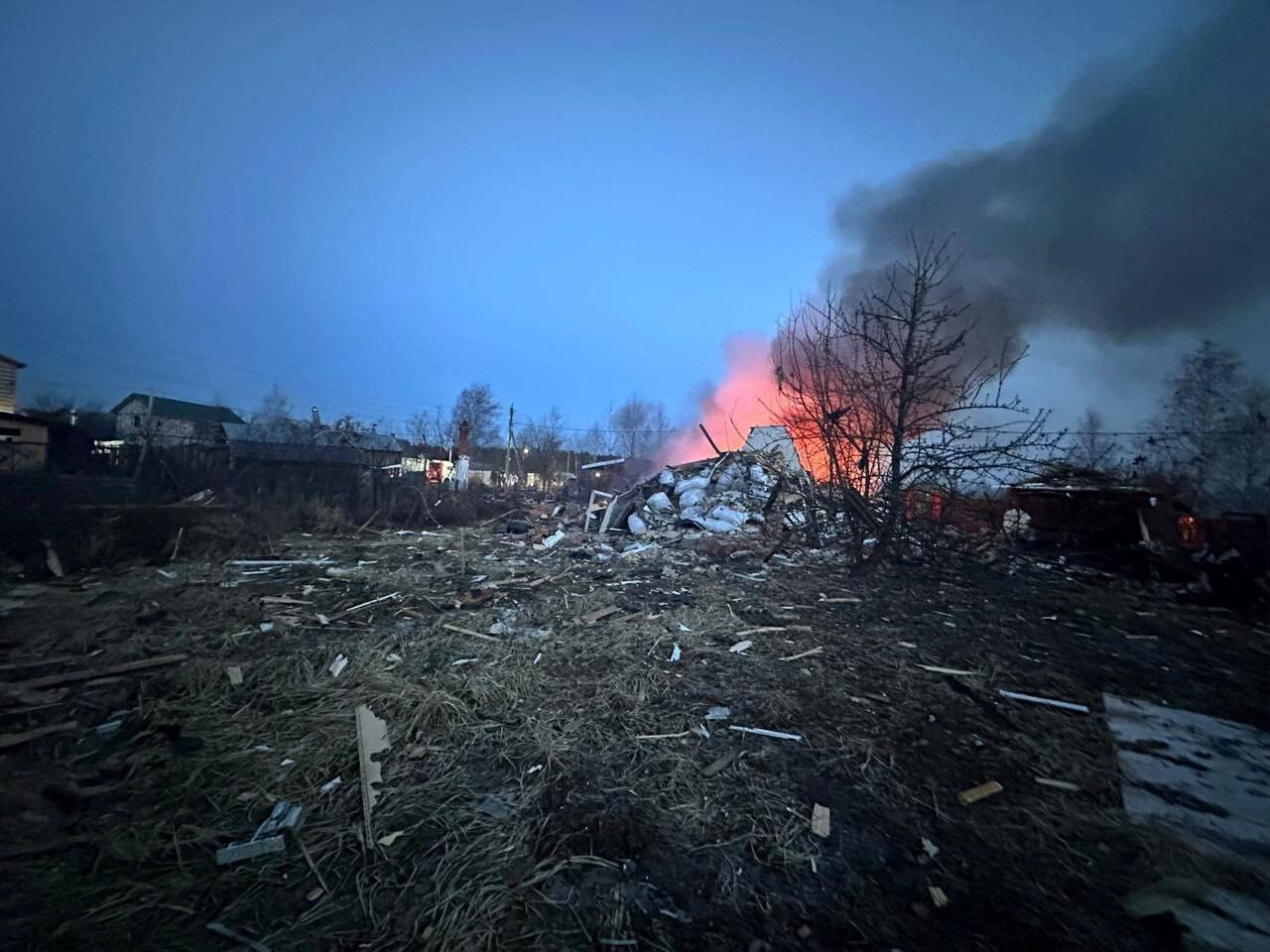 A charred grassy embankment cluttered with debris and a burnt skeleton of a tree in front of an orange plume of smoke