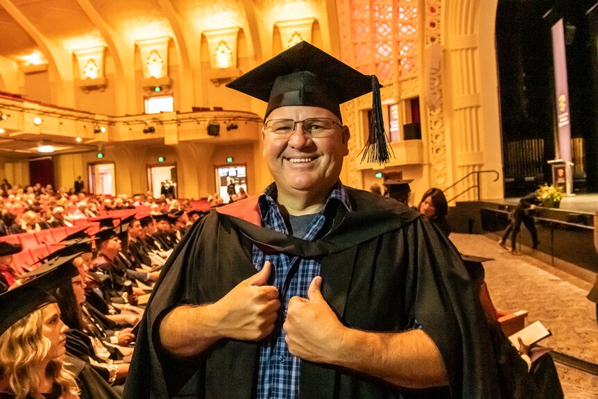 A man in a university cap and gown smiles at the camera giving two thumbs up