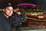 Night shot of Haidarr smiling while leaning on camera pointed at stadium lit up by pink lights.