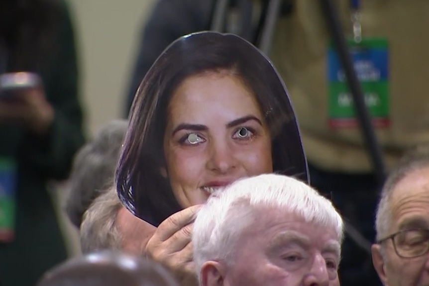 A man holds up a mask of Moira Deeming's face in a conference room.