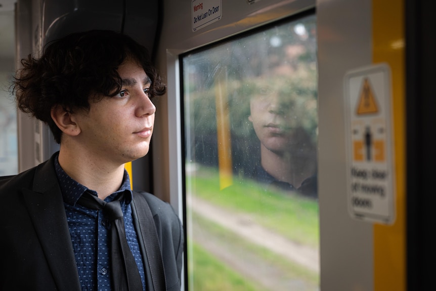 A young man looks out of a train window with a neutral expression, his face is reflected in the glass.