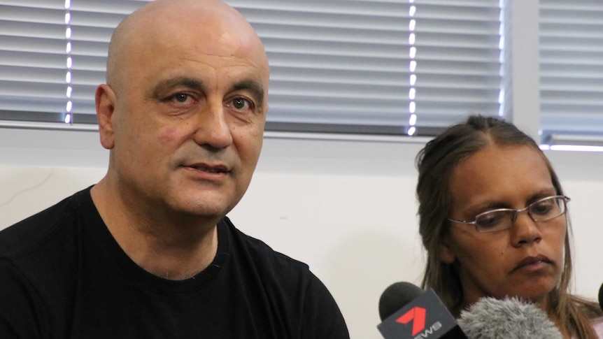 A man in a black t-shirt sits next to a woman wearing glasses in front of a set of microphones.