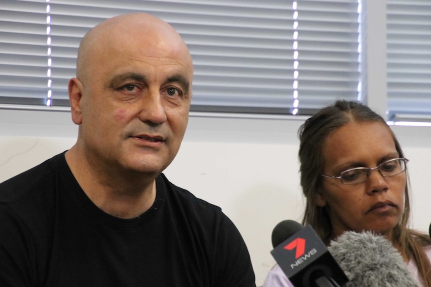 A man in a black t-shirt sits next to a woman wearing glasses in front of a set of microphones.