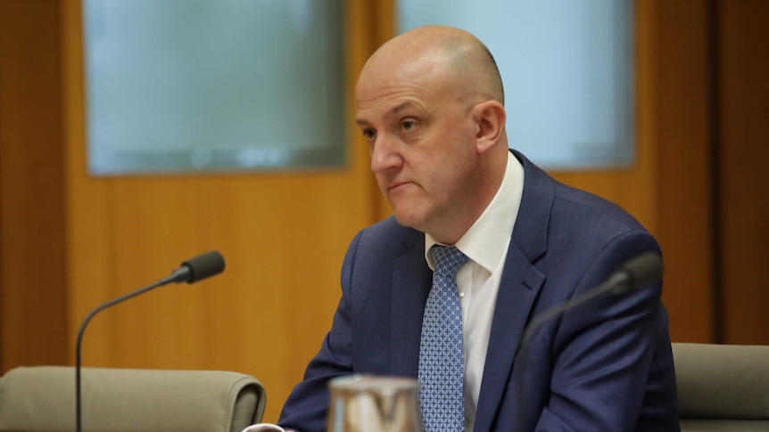 Mike Burgess sits at a table inside a committee room giving evidence to Parliament