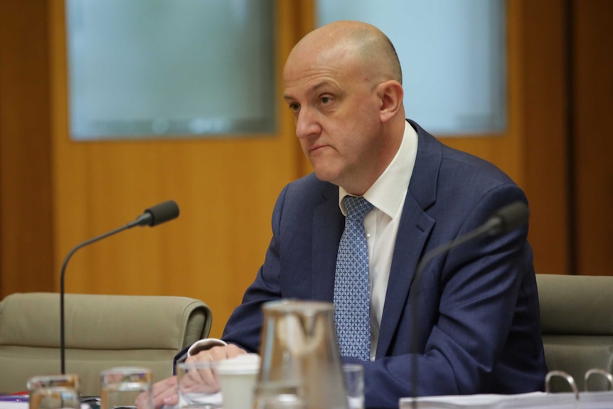 Mike Burgess sits at a table inside a committee room giving evidence to Parliament