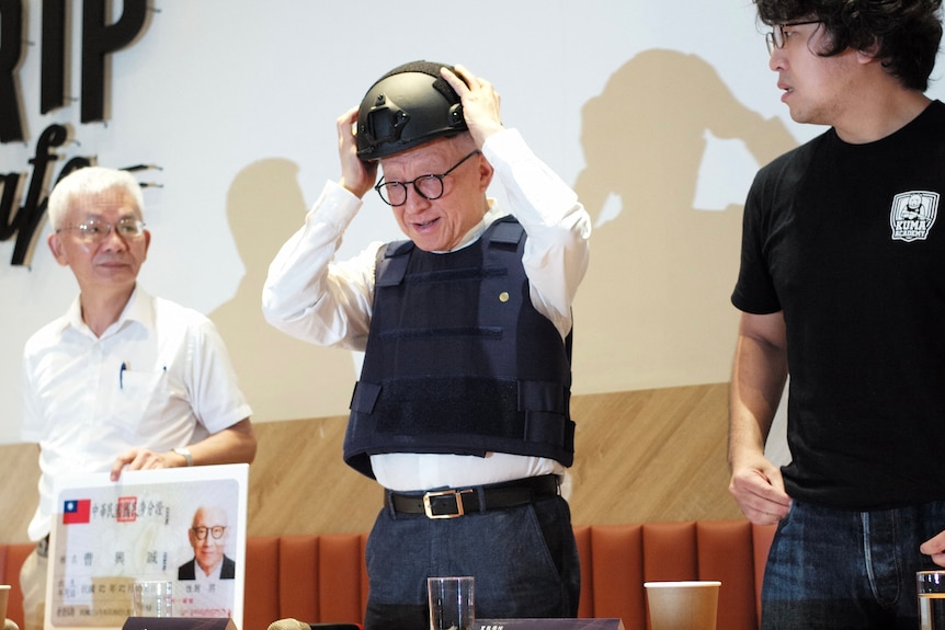 An older man wearing a blue bulletproof vest puts on a tactical helmet while smiling