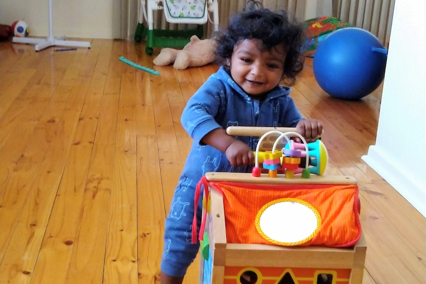 A young boy with a mop of black curls smiles as he plays with a toy while sitting on a timber floor.