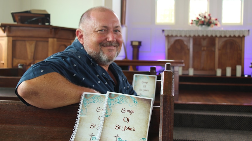 A man smiles at the camera sitting in a church pew with the altar behind him.