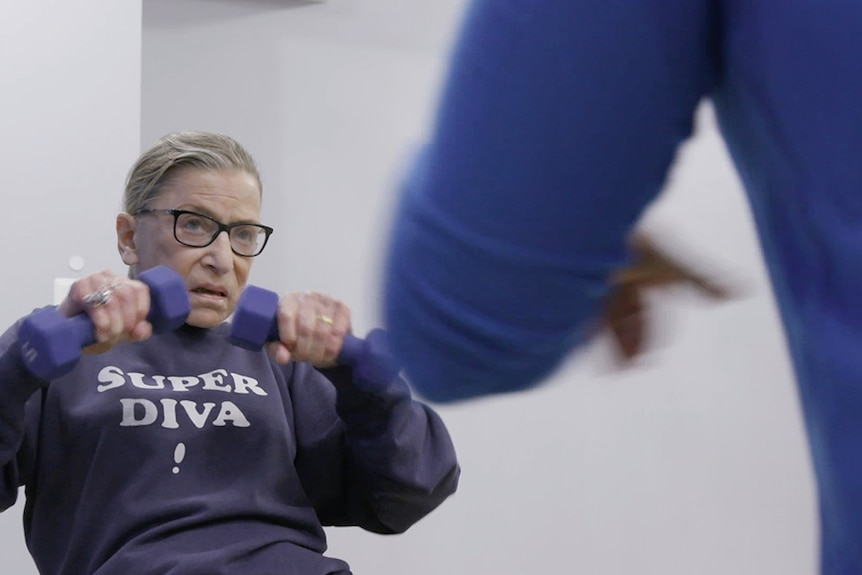 Colour still image from 2018 documentary RBG of Ruth Bader Ginsburg working out with dumbbells and a jumper.