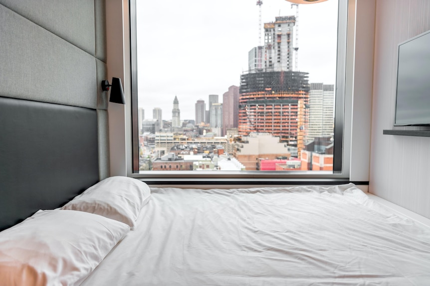 A small room with a big window with a view of a city with a bed filling the entire space with white linen