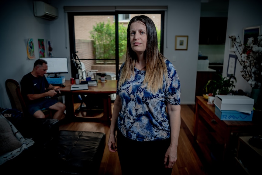 A woman with long brown hair standing in a living room while a man sits down at a table in the background