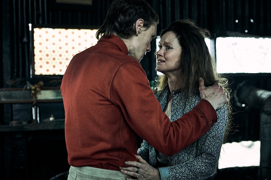 A man in red shirt and crying woman with worried and scared expression hold onto each other in dimly lit metal shed.