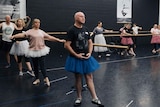 A man in the foreground wearing a blue ballet tutu taking a ballet lesson in a studio with others. 