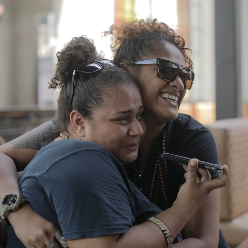 Two women hugging, one listening on a phone.