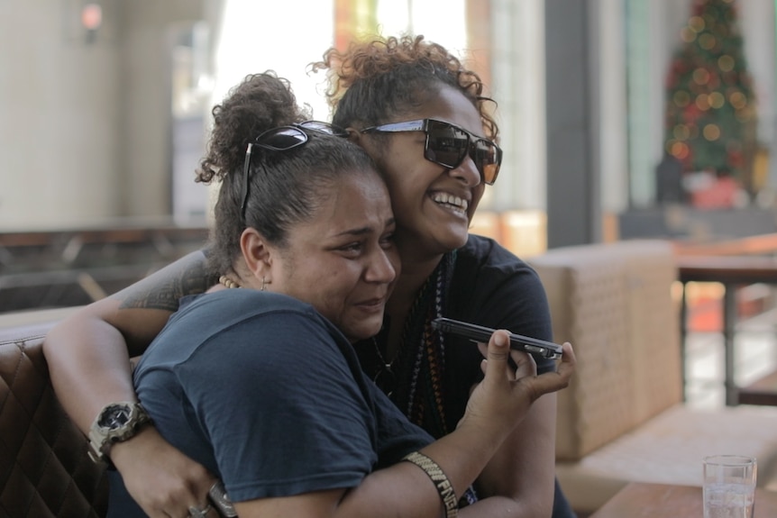 Two women hugging, one listening on a phone.