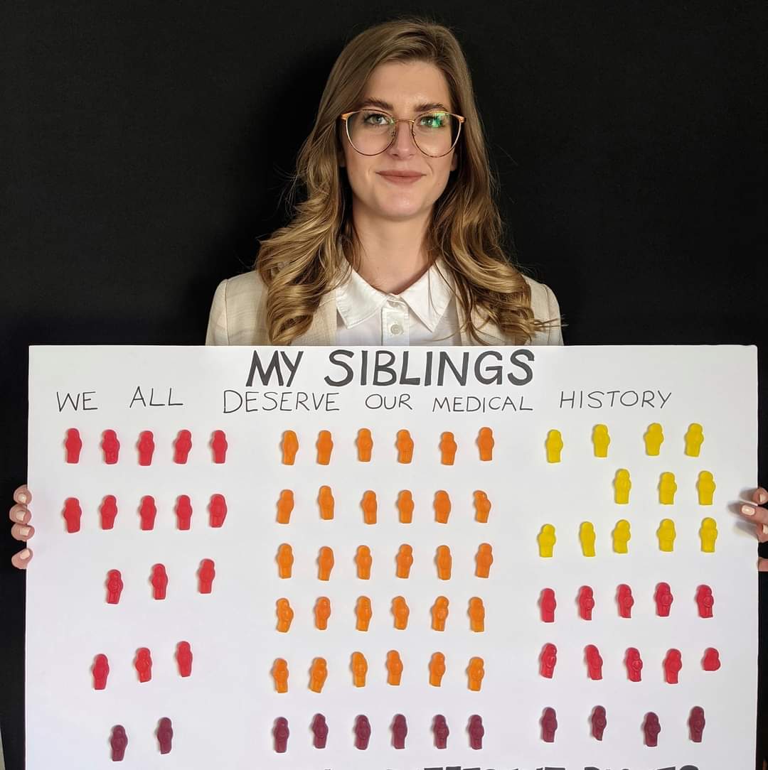 A woman with long hair and large round glasses holds a sign decorated with jelly babies which says 'My siblings...'