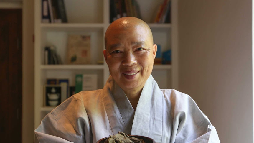 A woman with a shaved head and grey robe holds a plate of lotus-leaf wrapped dumplings, smiling.