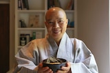 A woman with a shaved head and grey robe holds a plate of lotus-leaf wrapped dumplings, smiling.
