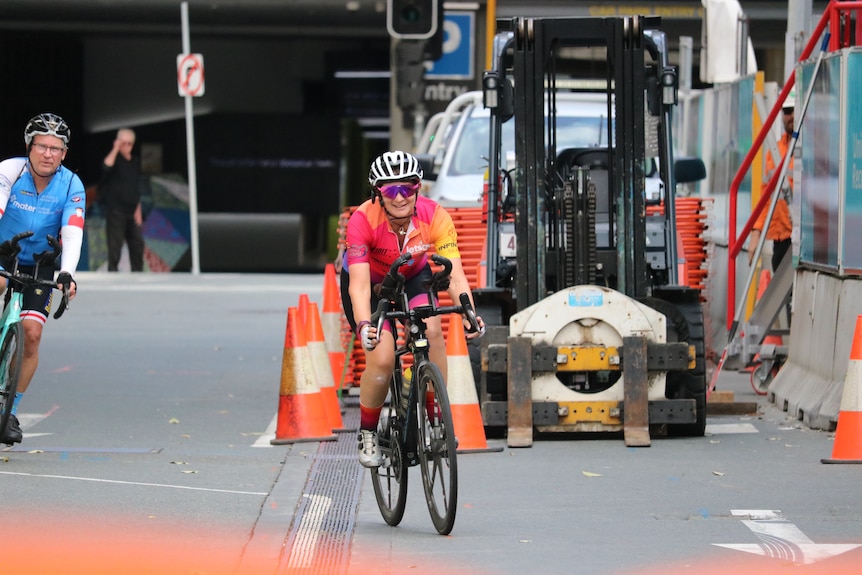 A woman cycles down bitumen, with construction work behind her.