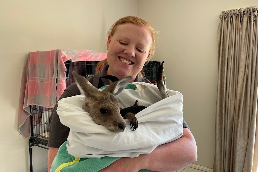 A smiling woman standing holding a small joey in a fabric pouch.