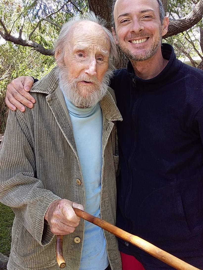 Peter Nicholas holds his walking stick towards the camera while Antonio Malevitsis puts his arm around him.