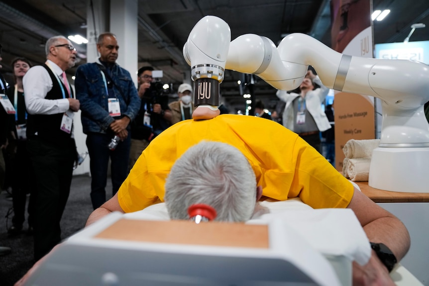 A close up near the head of a man lying on a bed and receiving a back massage from a robotic arm as people watch on