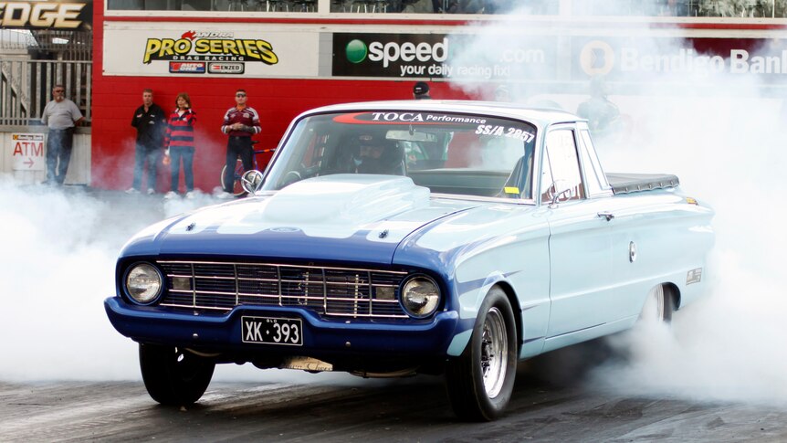 A classic muscle car lights it up on a track as handful of spectators watch its tyres going up in smoke.