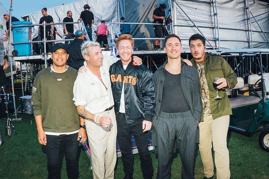 A group of men stand shoulder to shoulder smiling and laughing behind scaffolding of a stage 