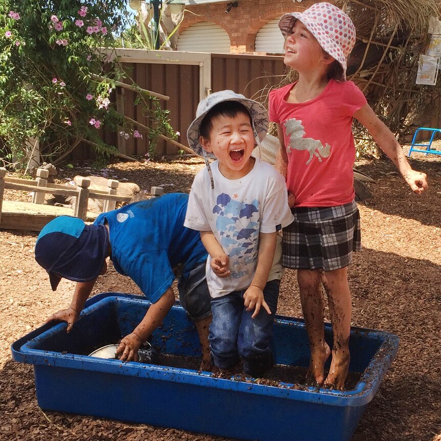 Three children standing in a bucket of mud.