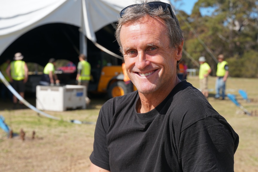 A man in a gray t-shirt smiling at the camera as people set up a large marquee tent in the background