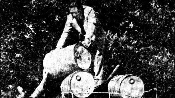a grainy black and white image of one man on a car roof and the other below with drums