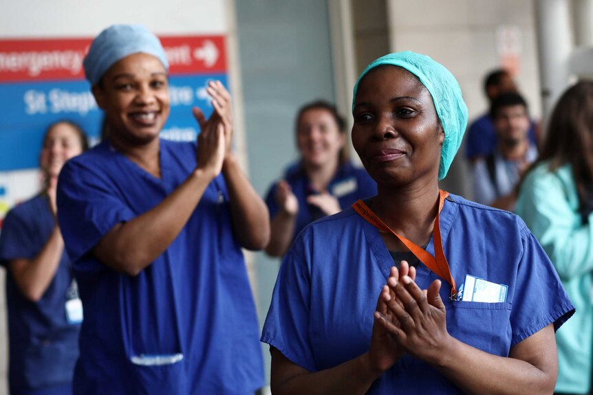 Two women wearing hairnets and scrubs clap outside a hospital