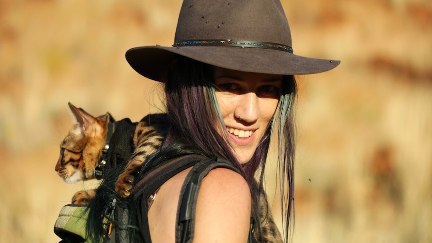 A woman with purple and green long hair wearing a hat carries a cat with dark brown spots in her backpack.
