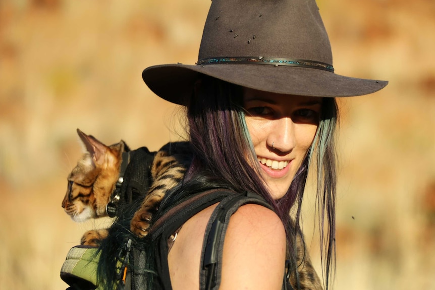 A woman with purple and green long hair wearing a hat carries a cat with dark brown spots in her backpack.