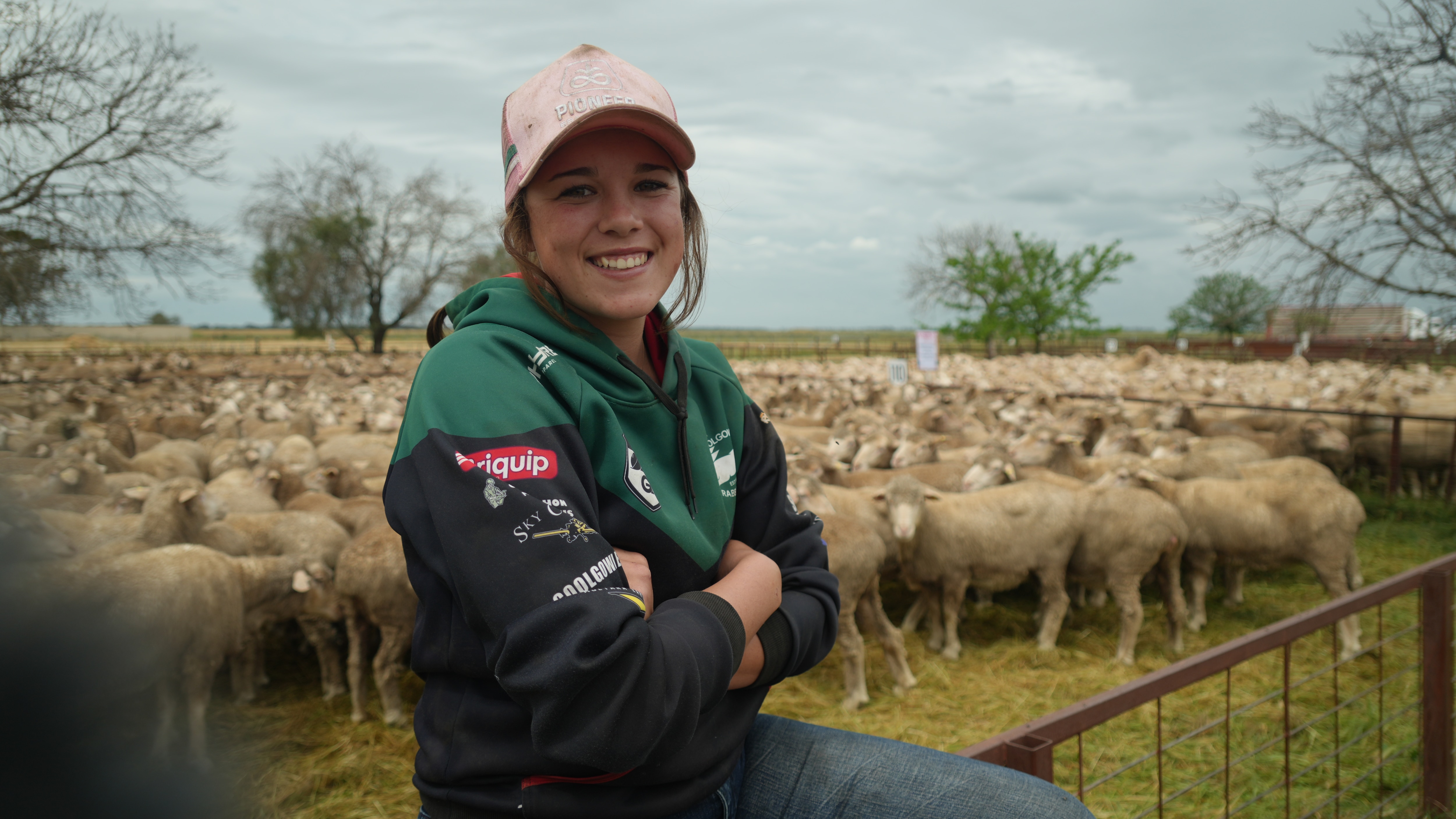 A young woman in a cap smiles at the camera with sheep in the background.