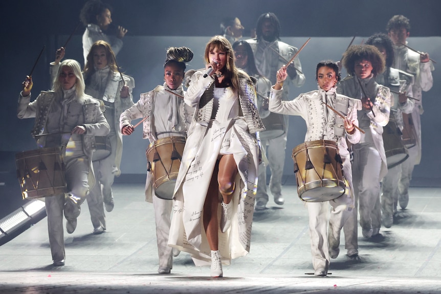 Taylor Swift on stage in a white dress and jacket with dancers drumming military drums behind her
