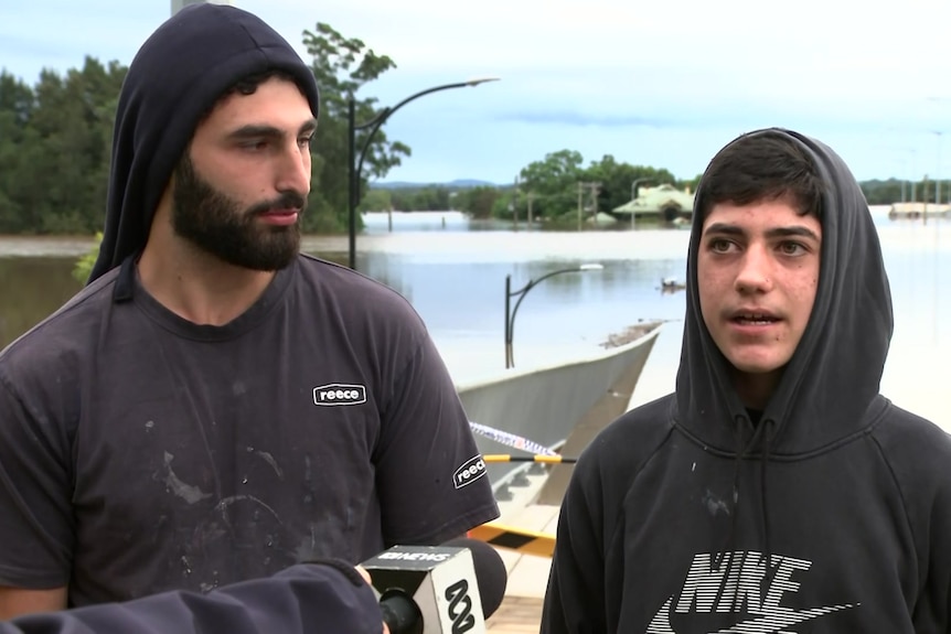 Two young men wearing hoodies look toward the camera with water behind them
