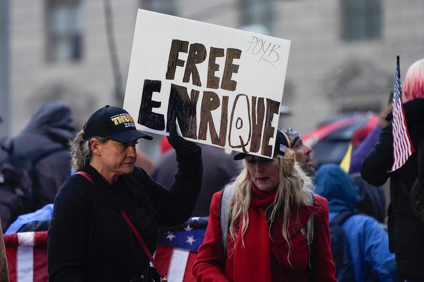 Woman in a cap holds a sign reading "Free Enrique" in a crowd.