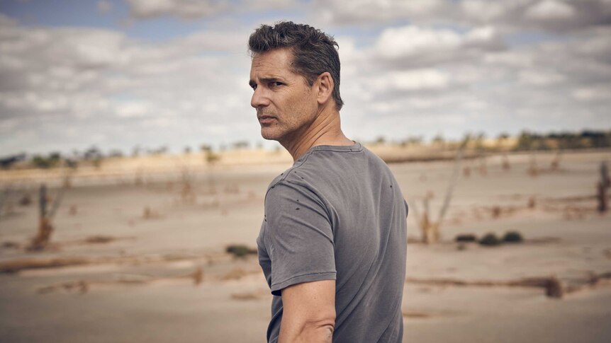 Eric Bana in a washed out dark grey t-shirt stands in a drought stricken plain side-on, and looks towards the camera.