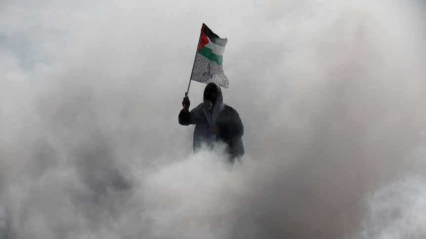 A hooded man is pictured waving a Palestinian flag. He is surrounded by smoke.