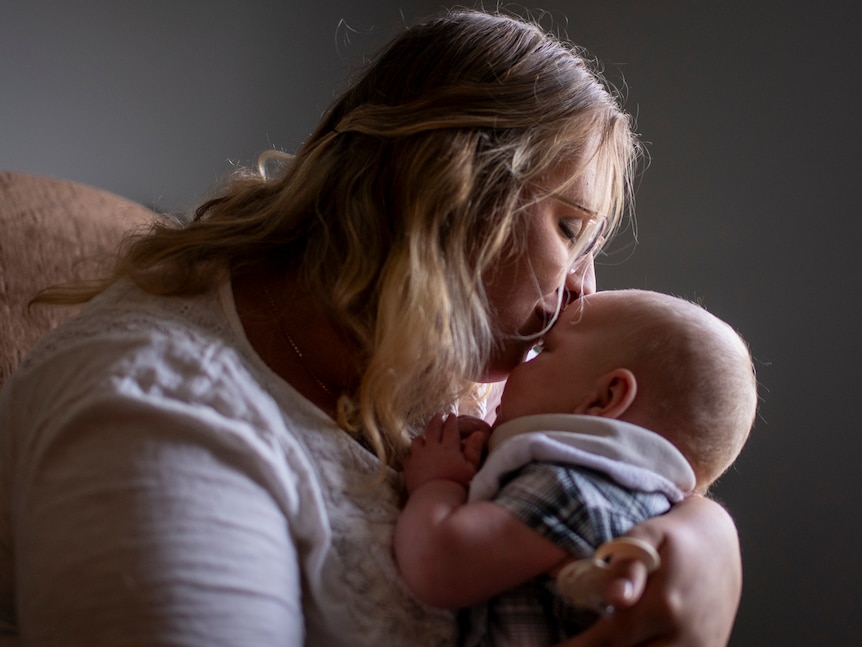 A mother kisses the forehead of her baby in soft window light.