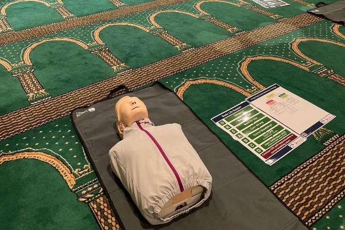 A dummy lies down inside a mosque while people kneel in the background
