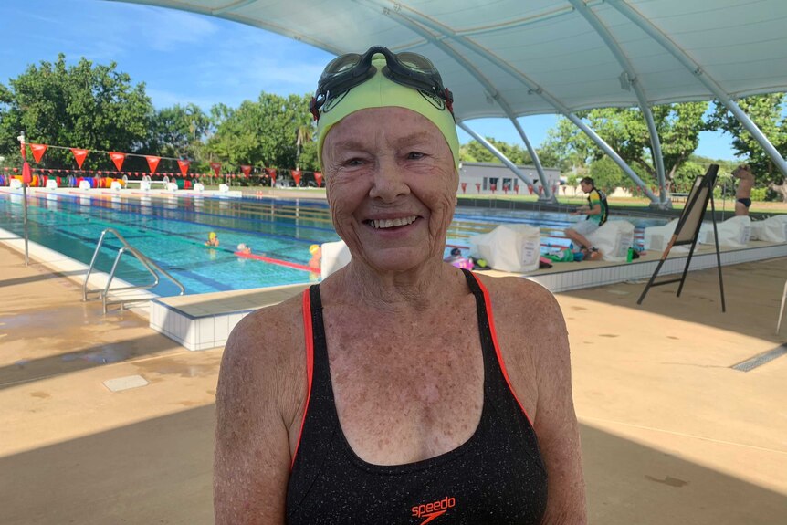 Anne Walker is wearing a black swim suit and a yellow swimming cap. She is smiling next to a pool.