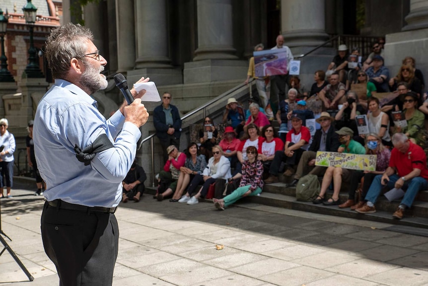 A man with a microphone and beard speaks to a crowd of people sitting on the steps at SA's Parliament House.