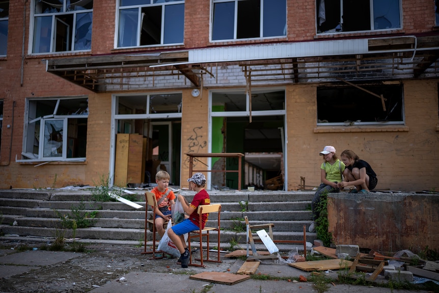 children sit outside a damaged school building in Ukraine