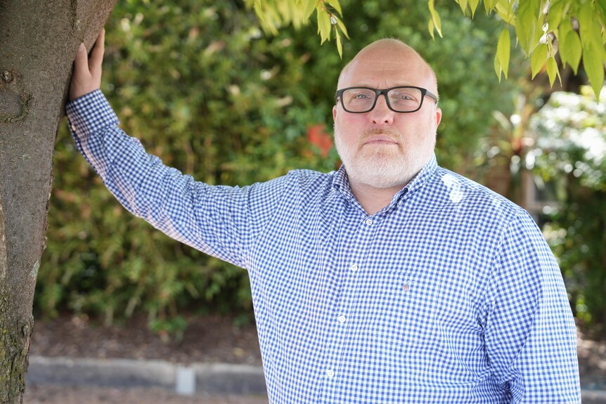 A man in a blue shirt looks at the camera and has his hand on a tree. He is wearing glasses. 